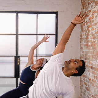 Two people practicing yoga in a studio with large windows and brick wall